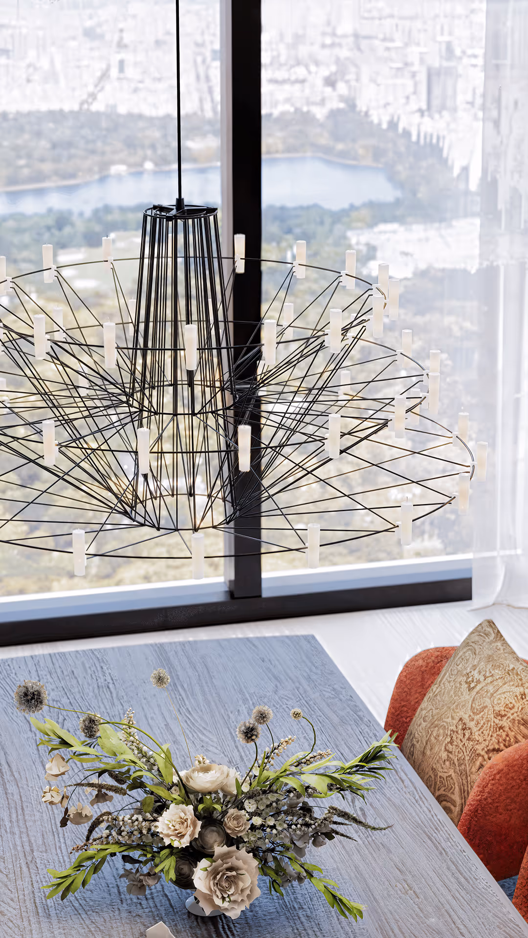 This space centers around a large circular artwork and a sculptural pendant light, aligned along a vertical axis to create a visually cohesive and artful composition. Together, they transform the dining area into a curated scene rather than a purely functional zone. A transparent glass table and ivory-toned, curved chairs emphasize openness and softness in form, while a deep green rug anchors the layout with subtle visual weight. The palette is grounded in warm neutrals, black, and natural wood, with a refined mix of leather, fabric, metal, and glass adding textural depth. A black Steinway & Sons grand piano becomes the artistic statement of the space, while lounge chairs with rich textures and patterned cushions complete the cozy, curated atmosphere.