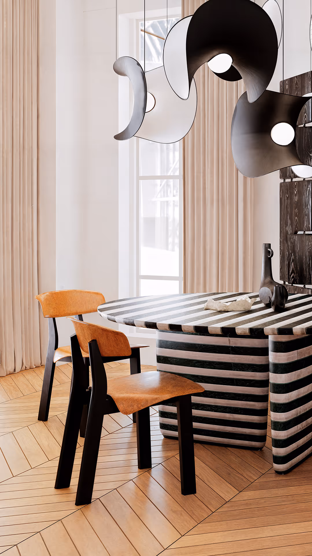 This dining space is curated around sculptural and curvilinear forms that create a dynamic, artistic environment. The centerpiece table features an oval top and asymmetric rounded legs, merging solidity with a sense of movement. Its black-and-white striped finish adds both rhythm and graphic clarity. The dining chairs complement this with softly contoured backs and seats that follow the body’s natural lines, contrasted by more structured, linear legs to maintain balance. Above, the pendant lighting draws from lunar phases, layering convex and concave shapes to add a poetic, sculptural canopy over the setting. Overall, the furniture composition communicates through form each piece contributing to a harmonious visual dialogue grounded in shape-driven storytelling.